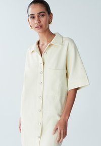 Šaty s krátkými rukávy a límečkem v světle žluté barvě, vyrobené z texturované látky, s knoflíky vpředu a náprsní kapsou na levé straně.