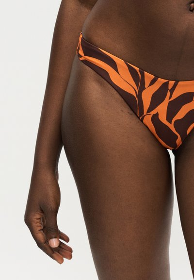 Άτομο με σκούρο δέρμα που φορά πορτοκαλί και καφέ μπικίνι με ρίγες ζέβρας, με χαλαρό αριστερό χέρι και χέρι ορατό σε λευκό φόντο.