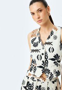 Chaleco sin mangas hecho de tela ligera texturizada, con patrones florales en negro y marrón, escote en V y botones delanteros para abrochar.
