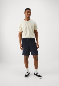 Licht beige T-shirt met korte mouwen, gecombineerd met marineblauwe shorts met een trekkoord. Het model draagt witte sokken en zwarte sneakers. Effen achtergrond.