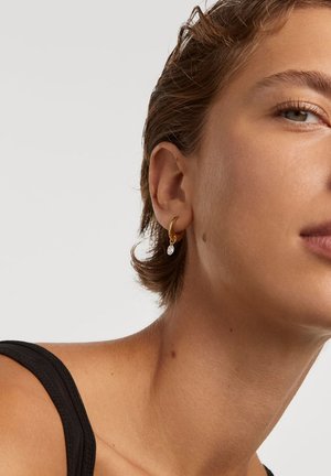 Guld hoopsørering med en lille, klar krystal dråbe vedhæng. Designet er enkelt, med en glat metallisk finish og let glans.