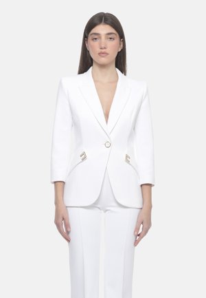 Donna che indossa un blazer bianco aderente con bottoni dorati e pantaloni bianchi abbinati, in piedi davanti a uno sfondo chiaro uniforme.