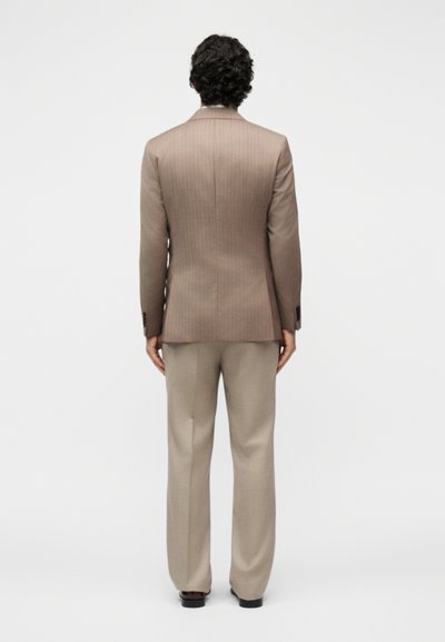 Άνδρας με σγουρά σκούρα μαλλιά φοράει μπεζ σακάκι με ρίγες και ανοιχτό μπεζ παντελόνι, στέκεται γυρισμένος πλάτη σε λευκό φόντο.
