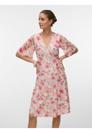 Naine kannab põlveni ulatuvat heleroosat lillelisi ümbermähitavat kleiti küünarvarte pikkuste varrukatega, seistes käed puusadel ühevärvilise tausta ees.