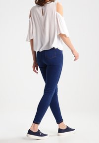 Blusa bianca a spalle scoperte con apertura a chiave sul retro, abbinata a jeans skinny blu scuro e sneakers navy. Tessuto morbido, vestibilità rilassata.