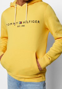 Sudadera amarilla con capucha hecha de mezcla de algodón. Presenta el logo "Tommy Hilfiger" con el año de fundación, bolsillo frontal tipo canguro y capucha con cordón ajustable.