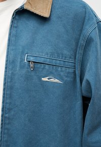 Μπλε μπουφάν με μπεζ γιακά από κοτλέ, λευκή κεντημένη λογότυπη, τσέπη στο στήθος με φερμουάρ, φοράται πάνω από λευκό πουκάμισο.