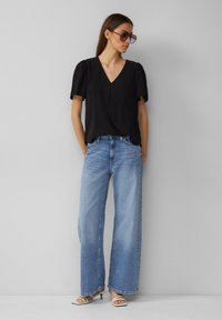 Blouse noire à col en V avec des manches bouffantes, associée à un jean large bleu clair et des sandales à talons blancs. Design minimaliste, tissu lisse.