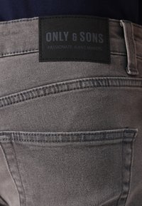 Sive traperice s crnom kožnom etiketom s natpisom "ONLY & SONS" i "PASIONIRANI PROIZVOĐAČI TRAPERICA" uz šivene džepove.