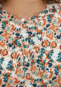 Blusa in tessuto floreale con macchie arancioni, turchesi e blu navy su sfondo bianco. Chiusura frontale con bottoni e scollo rotondo.