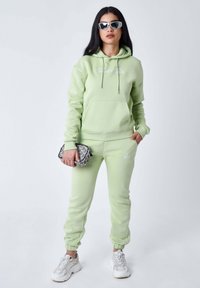 Sudadera verde y joggers a juego en tejido suave, con un bolsillo frontal y logo bordado. Accesorios incluyen un bolso de diseño y gafas de sol.