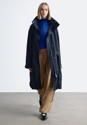 Langer schwarzer Regenmantel mit hohem Kragen und Reißverschluss, getragen über einem blauen Oberteil und beigen weiten Hosen. Glatter Stoff mit lässiger Passform.