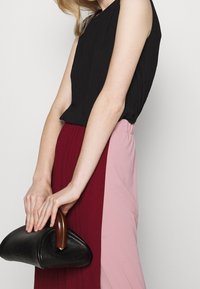 Person med en svart ärmlös topp och en färgblockerad kjol som håller i en svart läderhandväska med ett böjt trähandtag.