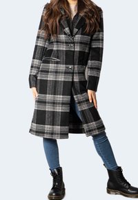 Žena oblečená v dlouhém černobílém kostkovaném kabátu, modrých džínách a černých šněrovacích botách, stojící s roztaženýma nohama na jednobarevném pozadí.