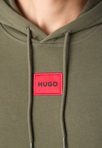 Gros plan d'un sweat à capuche vert olive avec cordons et un patch rectangulaire rouge portant la mention HUGO au centre devant.