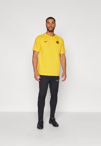 Mand står i en gul kortærmet Nike skjorte med logo, sorte Nike sweatpants og sorte sneakers mod en ensfarvet baggrund.
