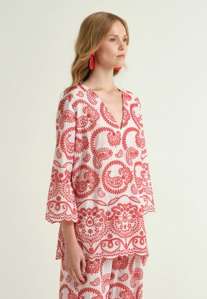 Femme portant un ensemble tunique blanche et pantalon avec broderie rouge paisley et boucles d'oreilles rouges, debout devant un fond clair uni.