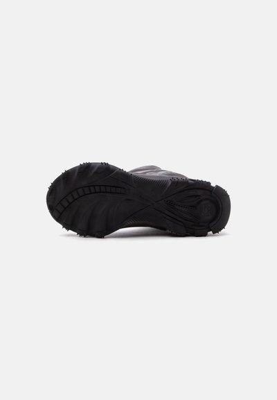 Semelle d’une chaussure de sport noire avec des crampons profonds et de petites pointes en caoutchouc pour l’adhérence, présentée sur un fond clair uni.