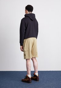 Cropped black hoodie with a hood, khaki shorts with side pockets, and brown boots with orange accents. Smooth texture and relaxed fit.
