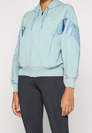 Persona que lleva una sudadera con capucha azul claro con cremallera, detalles verdes y paneles azules brillantes en las mangas, combinado con leggings negros.