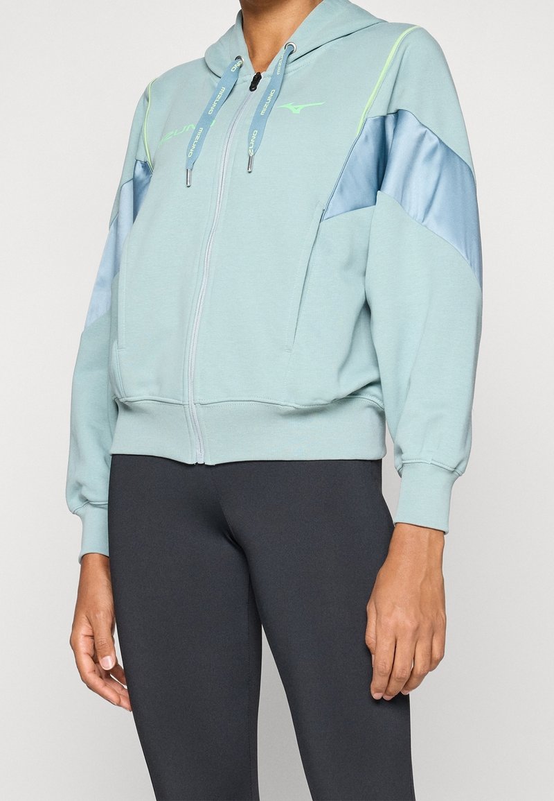Personne portant un sweat à capuche zippé bleu clair avec des accents verts et des panneaux bleus brillants sur les manches, associé à des leggings noirs.