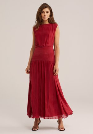 Kobieta w czerwoną, plisowaną, długą suknię bez rękawów i czarne sandały na obcasie, stojąca na jasnym, jednolitym tle.