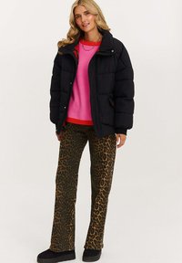 Blondýnka v černé péřové bundě, růžovém svetru, kalhotách s leopardím potiskem, černých botách a s korálkovým náhrdelníkem stojí před bílým pozadím.