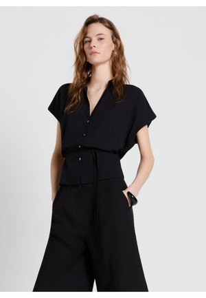 Femme aux cheveux ondulés portant un chemisier noir ample à boutons et un pantalon large noir, debout avec les mains partiellement dans les poches sur un fond gris.