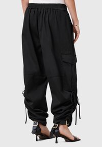 Pantalon cargo ample noir avec taille élastique et cordons aux poignets, porté avec des sandales à talons hauts noires à lanières, vu de dos.