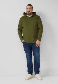Oliwkowa bluza z kapturem o teksturowanej tkaninie, z detalem szwu z przodu i logo. Połączona z ciemnoniebieskimi dżinsami i białymi trampkami.