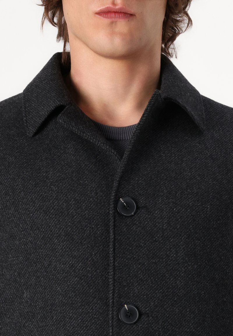 Mann in einem dunkelgrauen strukturierten Wollmantel mit breitem Kragen und zwei sichtbaren schwarzen Knöpfen, über einem dunklen gerippten Pullover.