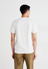 Hvit bomulls t-skjorte med korte ermer, ensfarget bak, rund hals; sammen med beige corduroy bukser med vertikale striper.
