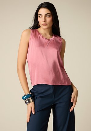 Kobieta w różowej satynowej bluzce bez rękawów, granatowych spodniach, z niebiesko-srebrnymi bransoletkami, z długimi ciemnymi włosami na beżowym tle.