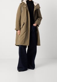 Parka khaki con cappuccio foderato in pelliccia color crema, zip frontale completa e chiusura con bottoni a pressione; abbinata a pantaloni in denim blu scuro larghi.
