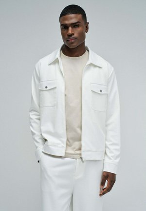 Junger Mann mit kurzen Haaren, der eine weiße Jacke mit Reißverschluss über einem beigen Hemd und weißen Hosen trägt, stehend vor einem schlichten grauen Hintergrund.