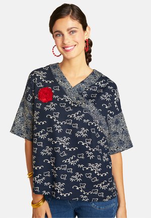 Frau lächelt, trägt ein marineblaues, kurzärmliges Top mit Blumenmuster, eine rote Blumenbrosche, rote Creolen und goldene Armreifen am Handgelenk.