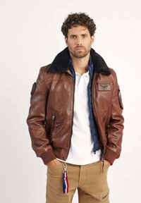 Veste bomber en cuir marron avec un col en fausse fourrure bleu marine, poches zippées et un patch sur la poitrine. Accompagnée d'un polo blanc et d'un pantalon kaki.