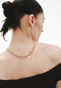 Collar de cadena de oro con grandes eslabones entrelazados y un pequeño colgante en forma de candado, sobre un hombro descubierto y una blusa negra.