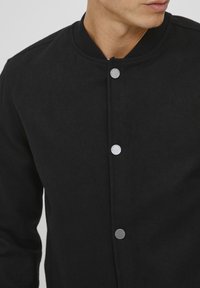 Svart bomberjakke med rund krage, teksturert stoff og sølvfargede trykknapper, med et minimalistisk design uten mønstre.
