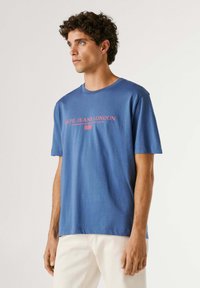 Jeune homme aux cheveux bouclés portant un t-shirt bleu Pepe Jeans London et un pantalon blanc, debout devant un fond clair uni.