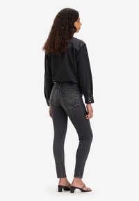 Jeans skinny de mezclilla negra con acentos de tachuelas plateadas en los bolsillos traseros, combinados con una camisa negra de corte holgado y sandalias de tacón negro.