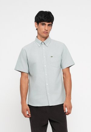 Chemise légère gris clair à manches courtes, avec col, poche poitrine unique et un petit logo vert. Tissu texturé, coupe slim.