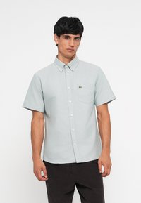 Light grey short-sleeve button-up shirt with a collar, single chest pocket, and a small green logo accent. Textured fabric, slim fit.