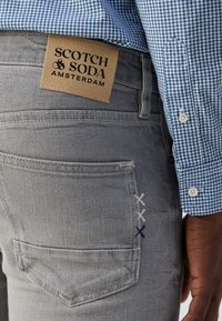 Šedé džíny z denimu s béžovou koženou nášivkou označenou "SCOTCH & SODA AMSTERDAM." Mají výrazné šití v okolí zadního kapsy.