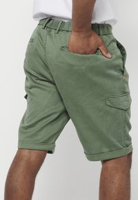 Muškarac u maslinasto zelenim cargo kratkim hlačama s rukom u stražnjem džepu, na glatkoj bijeloj pozadini.