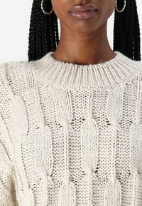 Gros plan sur une personne portant un pull en maille épaisse de couleur crème avec un motif feuille texturé et des boucles d'oreilles argentées en forme d'anneau.