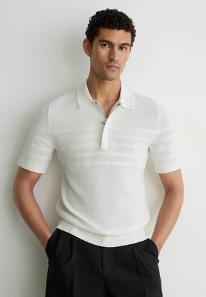 Mann trägt ein weißes kurzärmeliges Strick-Poloshirt mit strukturierten horizontalen Streifen, schwarze Hose, steht an einer weißen Wand.