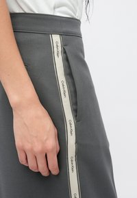 Γκρι φούστα με εφαρμοστή μέση, που διαθέτει πλαϊνή τσέπη και μπεζ ταινία διακοσμημένη με την επιγραφή «Calvin Klein» κατά μήκος της ραφής.