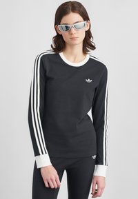 Žena oblečená v čiernom dlhom rukáve topu Adidas s bielymi pruhmi a logom, s nohavicami na telo, a reflexnými striebornými slnečnými okuliarmi, stojí pred bielym pozadím.