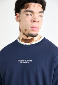 Tumši zila ilgi piedurkņu krekls ar baltu drukātu logo "STUDIO EDITION" un "ALPHA INDUSTRIES INC." Kaklasaites izgatavota no krāsainām pērlēm.